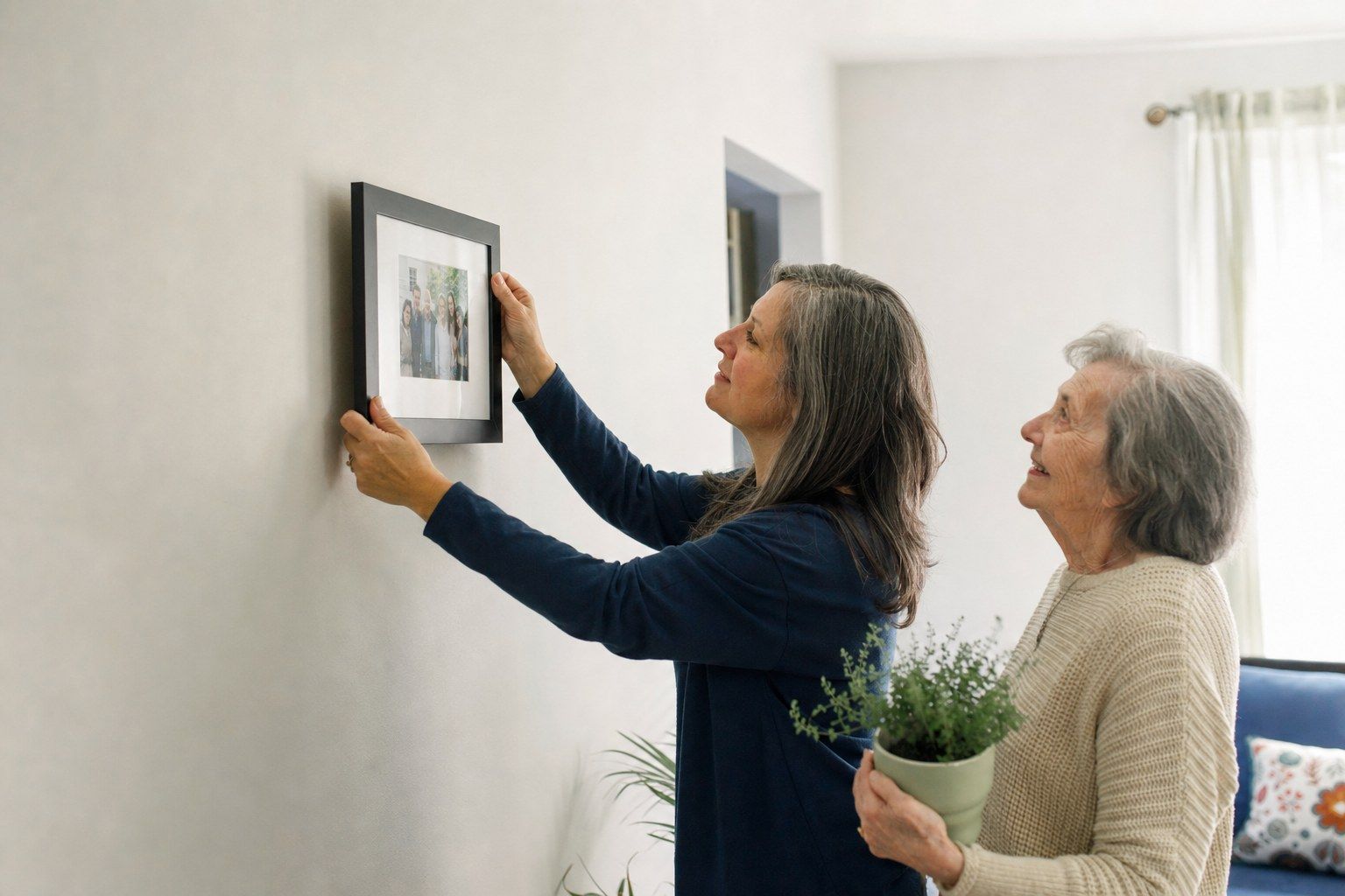 A woman hanging a framed family photograph on the wall of her mother’s new home while her mother watches