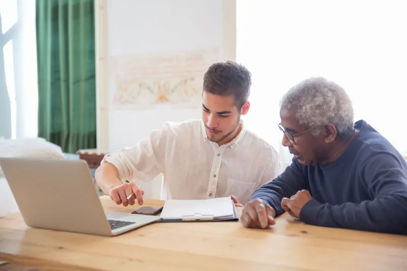 A younger man and an older man looking at paperwork and a laptop together at a table