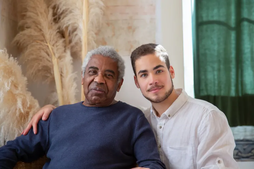 An older man sitting on a sofa with his adult grandson, both looking towards the camera